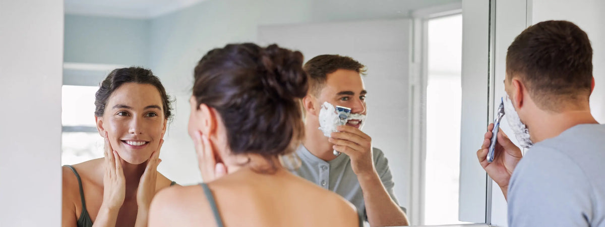 A man shaving in a bathroom while looking at his reflection, with a woman in the foreground and another man holding a phone visible in the mirror and side view.