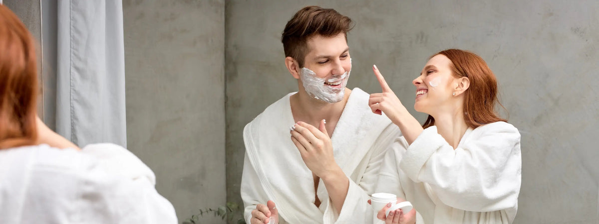 A man and a woman in white robes standing in front of a mirror, with the man shaving his face and the woman pointing at it. The man has shaving foam on his face.