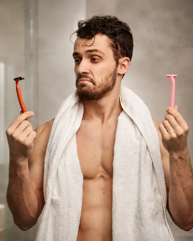 Man holding two razors, one red and one pink, in a bathroom setting.
